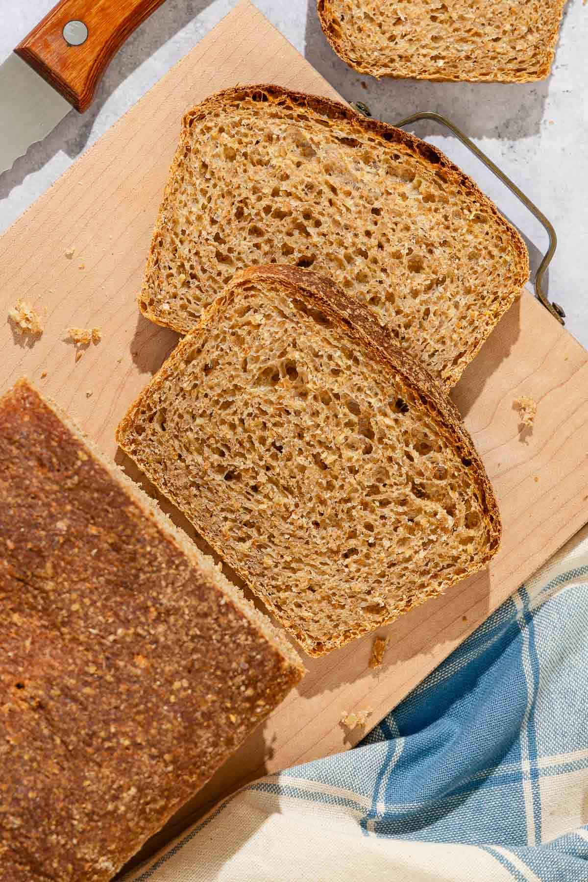 A partially sliced whole wheat bread loaf next to 2 slices of the bread on a cutting board.