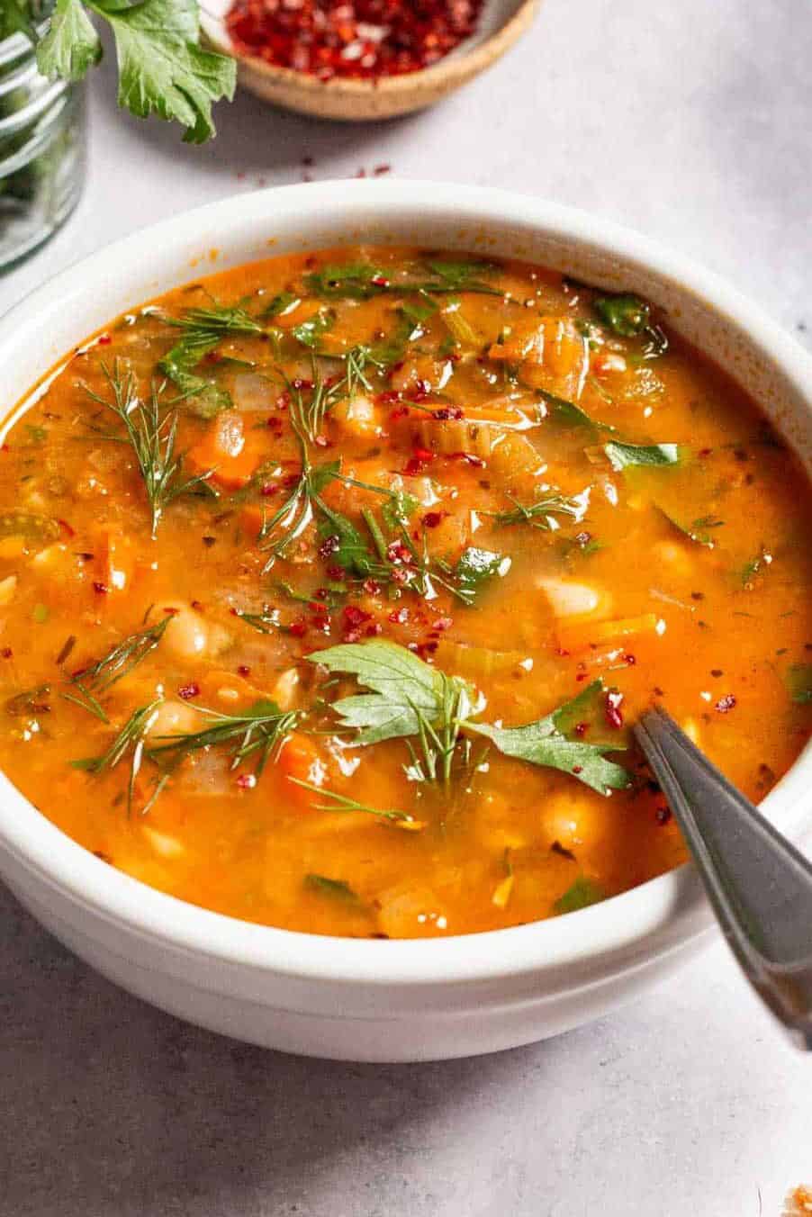 Mediterranean white bean soup in a bowl with a spoon next parsley, dill and a small bowl of aleppo pepper.