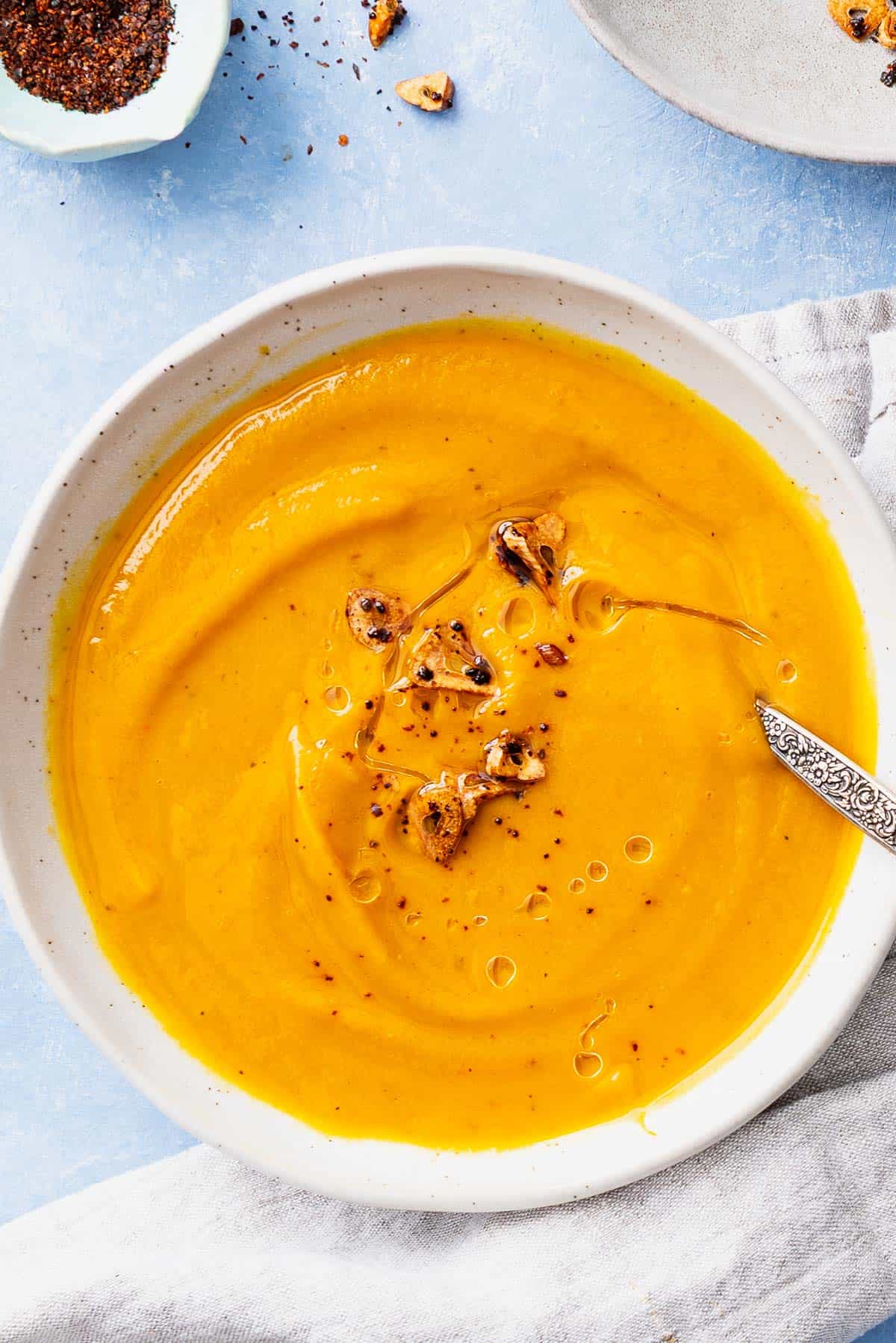 Pumpkin soup topped with the Aleppo pepper-garlic oil in a bowl with a spoon. Next to this is a cloth napkin, and a bowl of Aleppo pepper.
