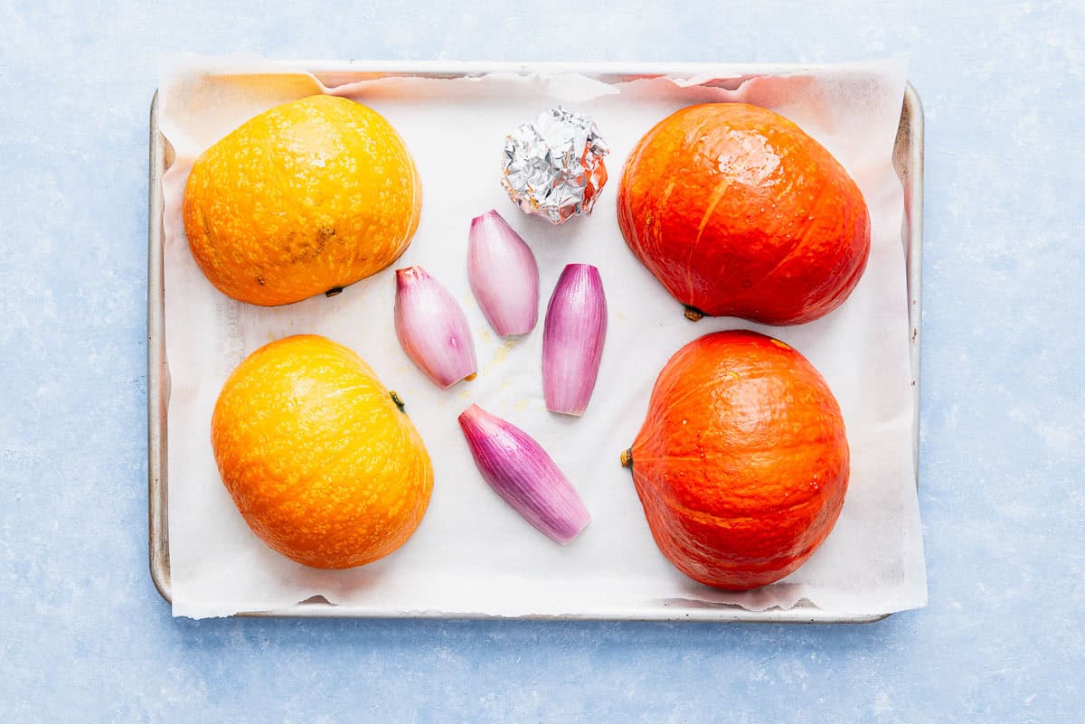 The uncooked halves of kuri, shallots and garlic wrapped in foil on a parchment lined sheet pan.