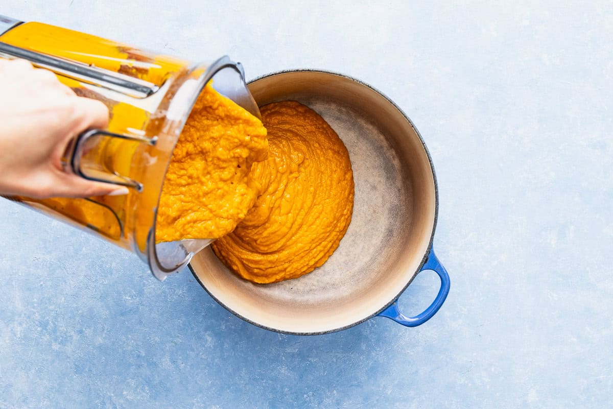 The pureed pumpkin soup being poured from the pitcher of a blender into a large pot.