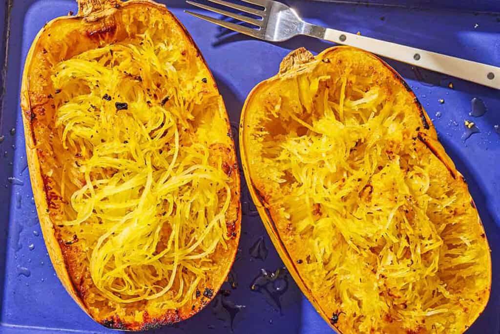 Two roasted spaghetti squash halves on a baking sheet with a fork after their flesh has been scraped.