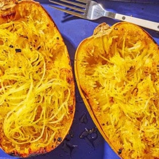 Two roasted spaghetti squash halves on a baking sheet with a fork after their flesh has been scraped.