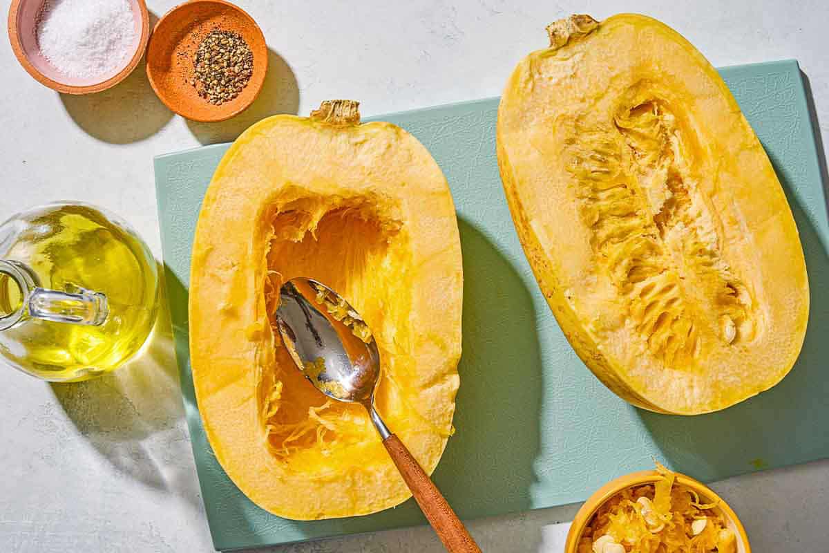 Two unbaked spaghetti squash halves on a cutting board, one with a spoon after its seeds have been scooped out. Next to these are bowls of salt and pepper, a bottle of olive oil, and a bowl of the spaghetti squash seeds.