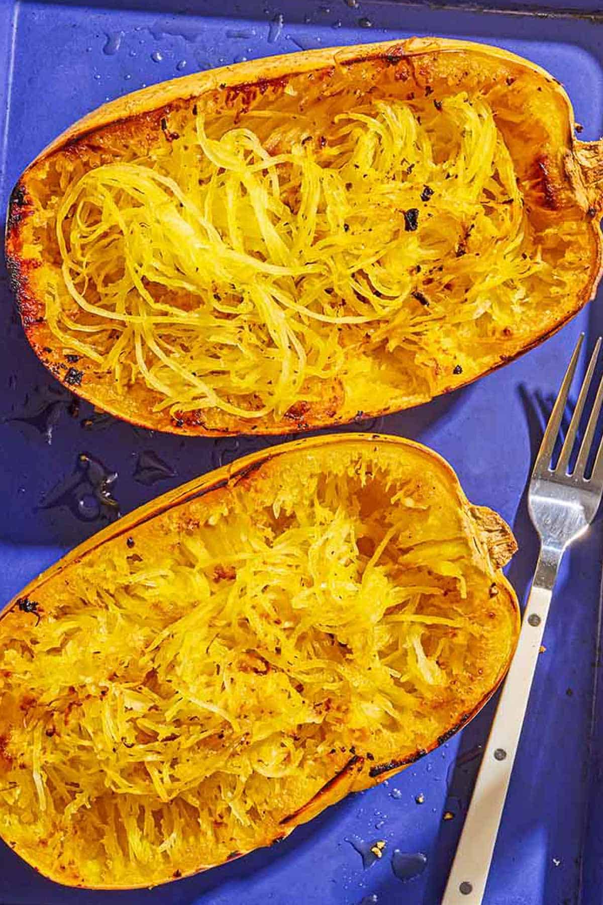 Two roasted spaghetti squash halves on a baking sheet with a fork after their flesh has been scraped.