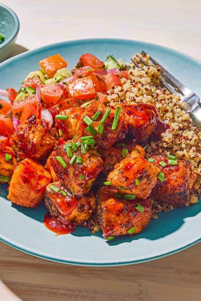A serving of the air fryer salmon bites topped with chopped chives on a plate with quinoa, tomato cucumber salad and a fork.