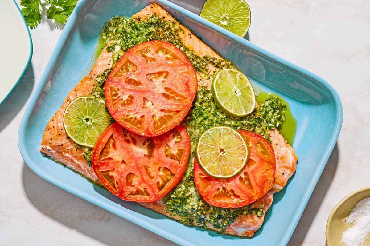 Uncooked cilantro lime salmon in a baking dish. Next to this is a bowl of salt, a lime wheel, a sprig of cilantro, and a plate.
