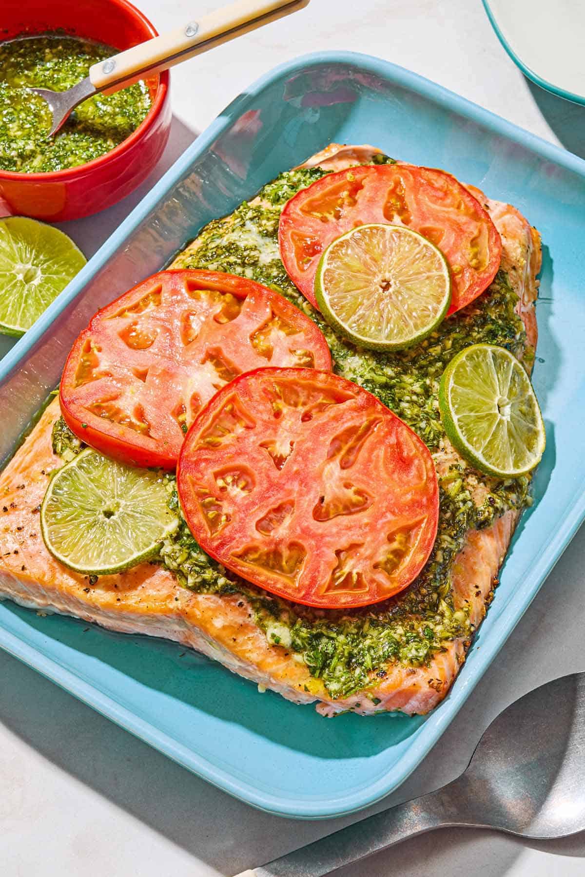 Uncooked cilantro lime salmon in a baking dish. Next to this is a serving spoon, a bowl of the cilantro sauce, and a lime half.