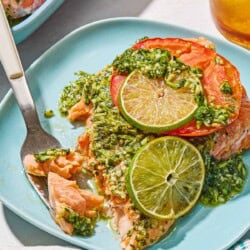 A serving of cilantro lime salmon on a plate with a fork. Next to this is a cloth napkin, a glass of sparkling eater and the rest of the cilantro lime salmon on a platter.