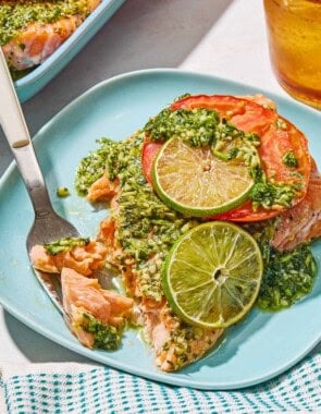 A serving of cilantro lime salmon on a plate with a fork. Next to this is a cloth napkin, a glass of sparkling eater and the rest of the cilantro lime salmon on a platter.