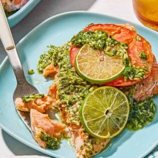 A serving of cilantro lime salmon on a plate with a fork. Next to this is a cloth napkin, a glass of sparkling eater and the rest of the cilantro lime salmon on a platter.