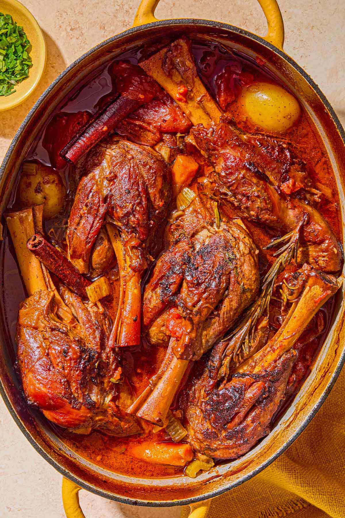 The braised lamb shanks in a large pot. Next to this is a bowl of chopped parsley.