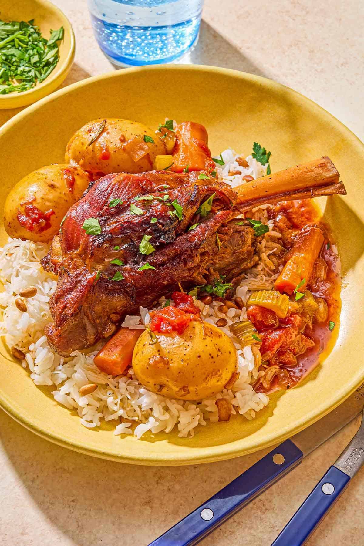 Braised lamb shanks with carrots and potatoes on a bed of rice in a bowl. Next to this is a bowl of chopped parsley, a glass of sparkling water, and utensils.