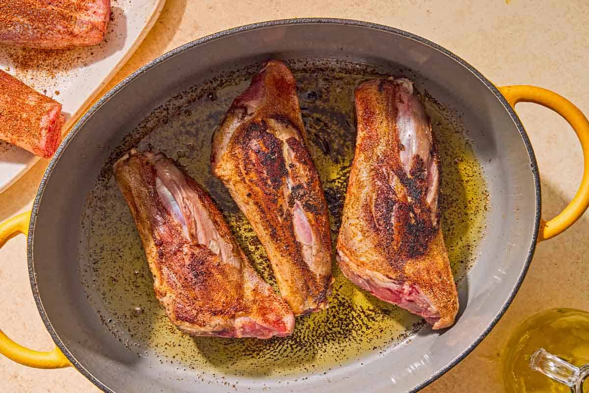 Three lamb shanks being browned in a large pot. Next to this is a plate with more seasoned shanks and a bottle of olive oil.