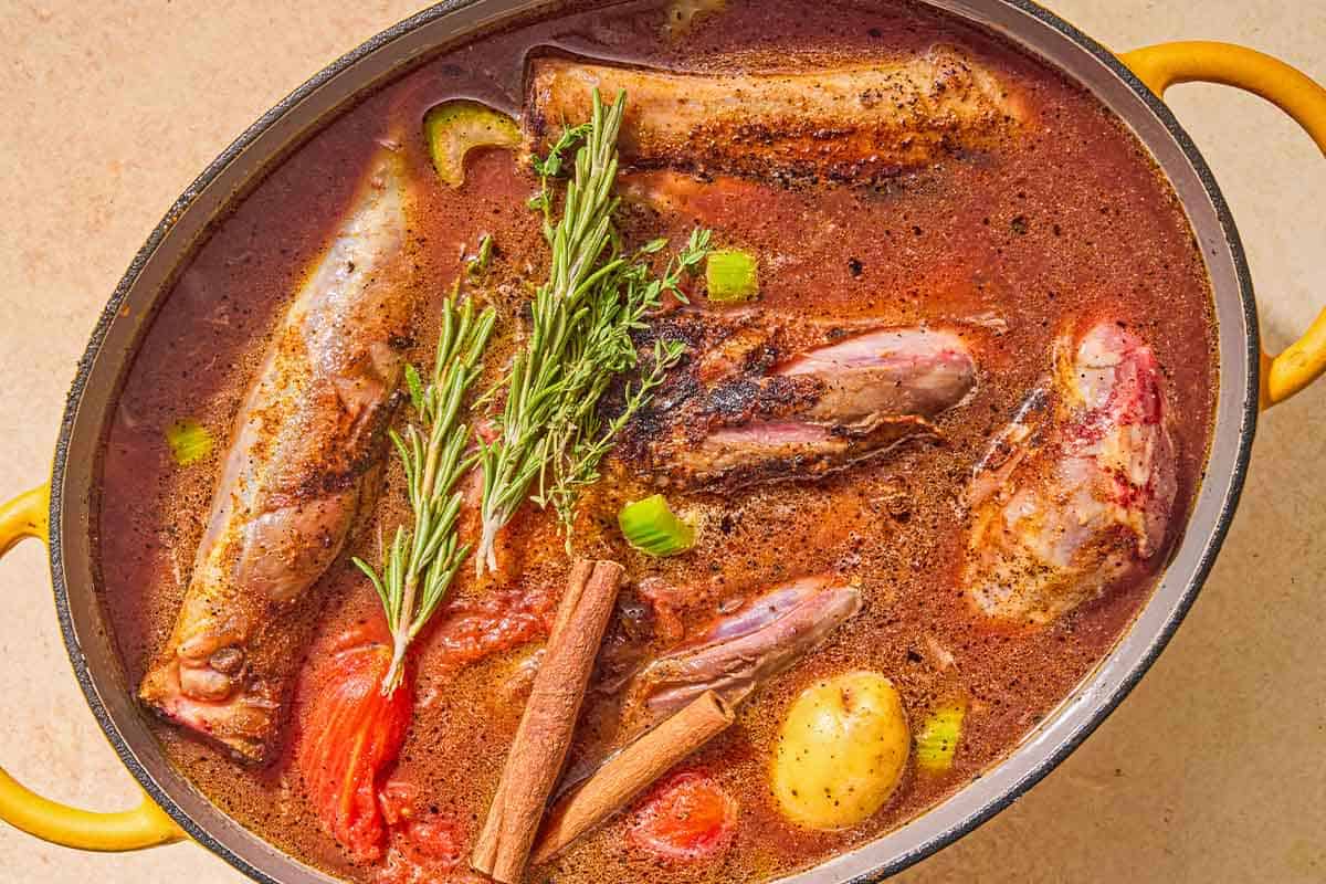 The ingredients for the braised lamb shanks in a large pot topped with fresh rosemary and thyme and cinnamon sticks.