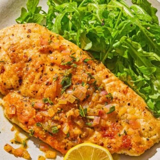 A serving of chicken scaloppine on a plate with an arugula salad and a lemon slice.