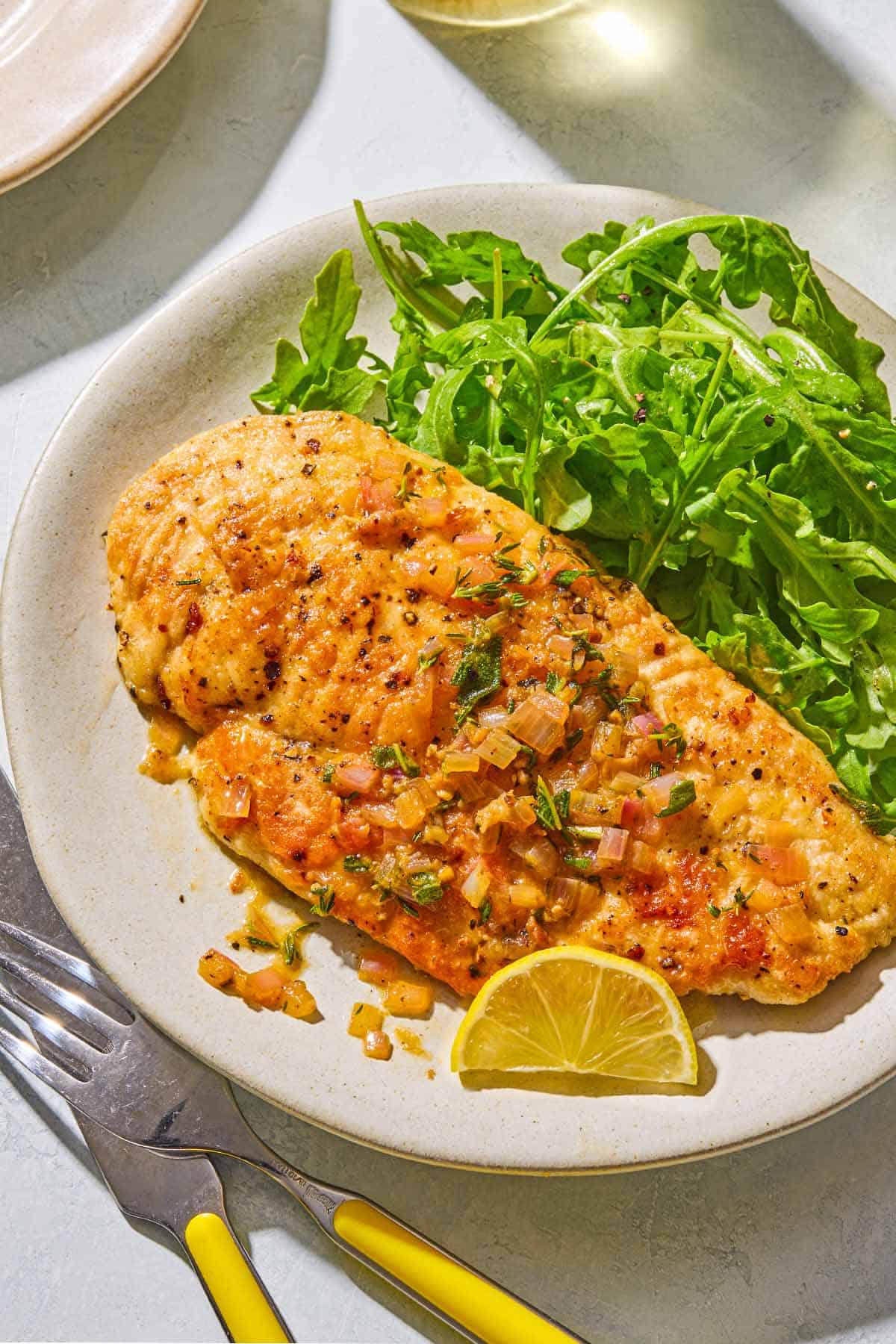 A serving of chicken scaloppine on a plate with an arugula salad and a lemon slice. Next to this is a knife and fork.