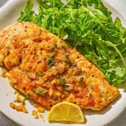 A serving of chicken scaloppine on a plate with an arugula salad and a lemon slice. Next to this is a knife and fork.