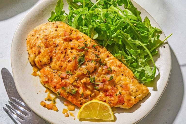 A serving of chicken scaloppine on a plate with an arugula salad and a lemon slice. Next to this is a knife and fork.