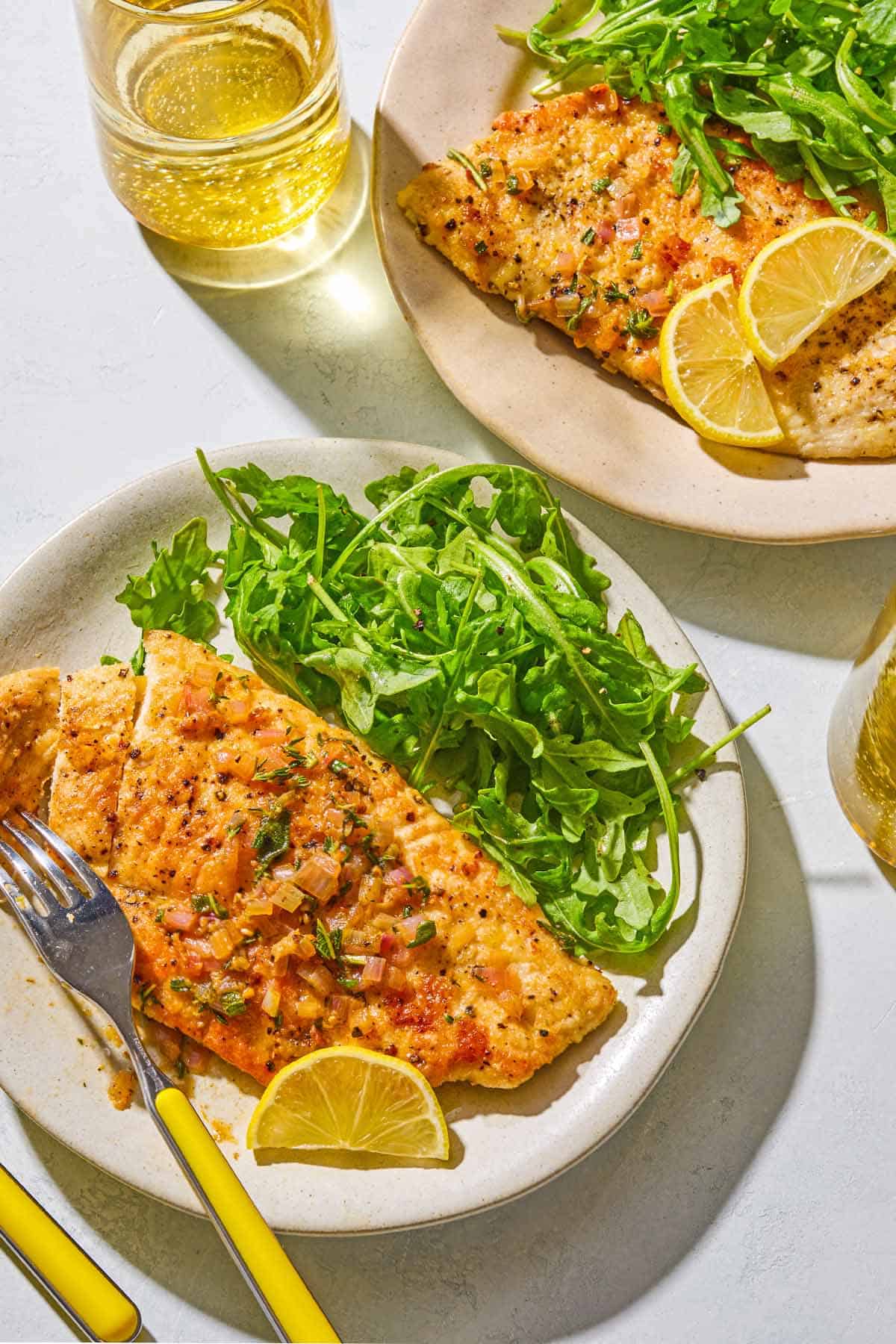 A serving of chicken scaloppine on a plate with an arugula salad, a lemon slice and a fork. Next to this is another serving of the chicken scaloppine and salad on a plate, and a glass of sparking water.
