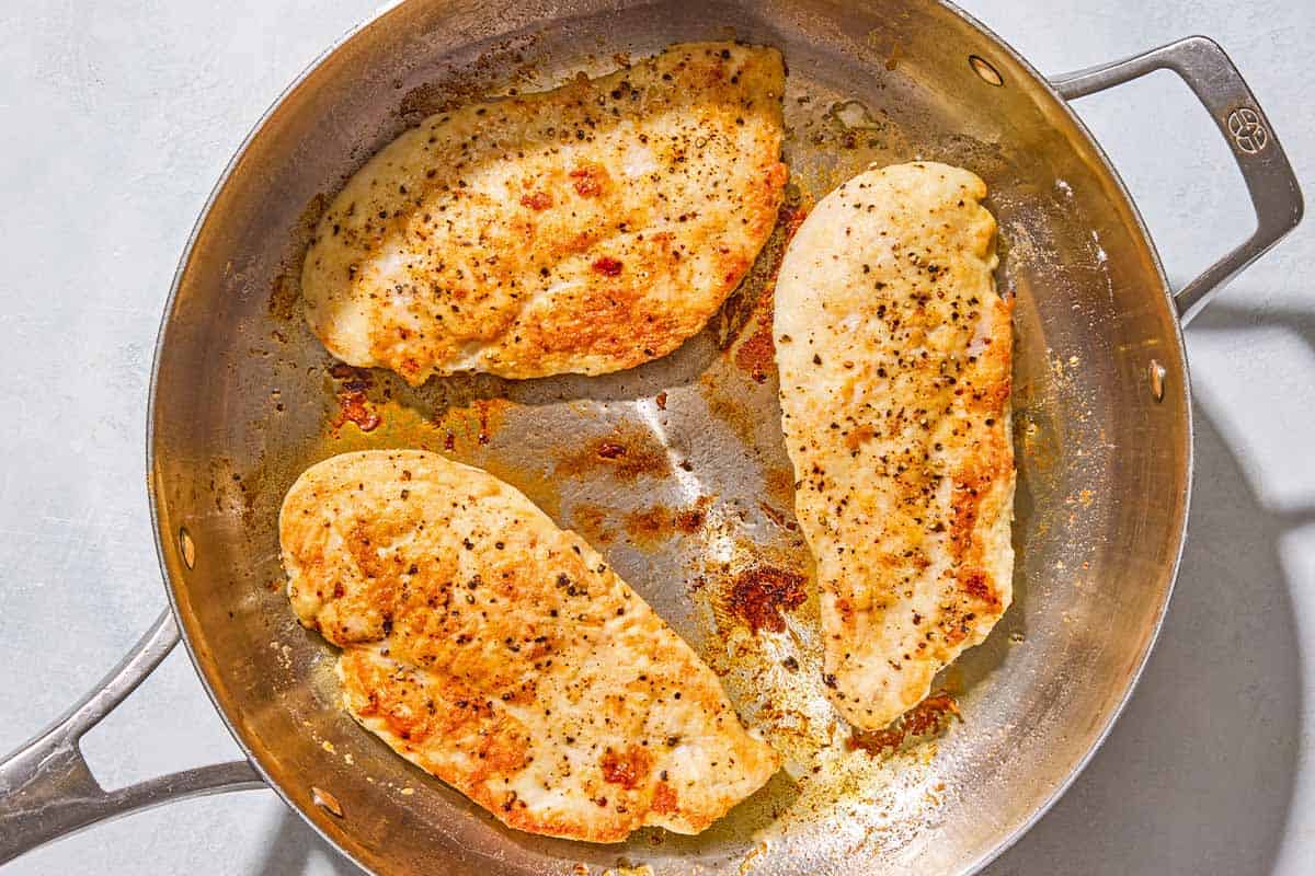 Three chicken breast cutlets being browned in a skillet.
