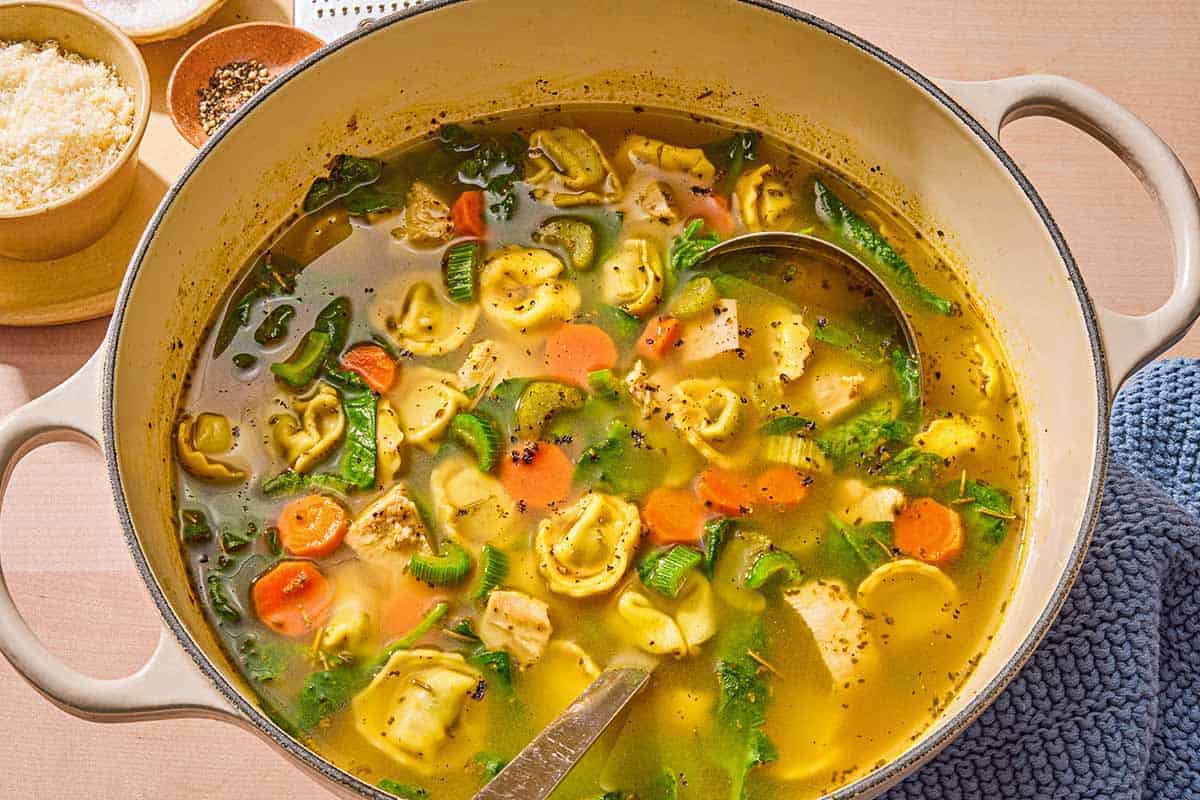 The tortellini soup in a large pot with a ladle. Next to this are bowls of grated Parmigiano Reggiano cheese and pepper.