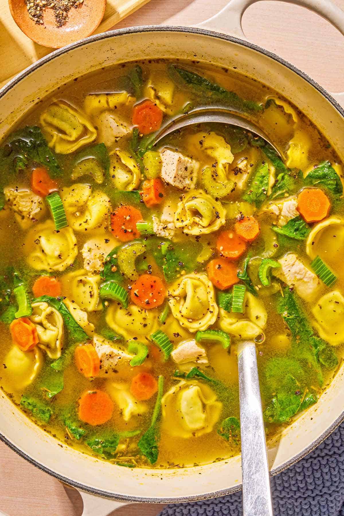 The tortellini soup in a large pot with a ladle next to a small bowl of pepper.