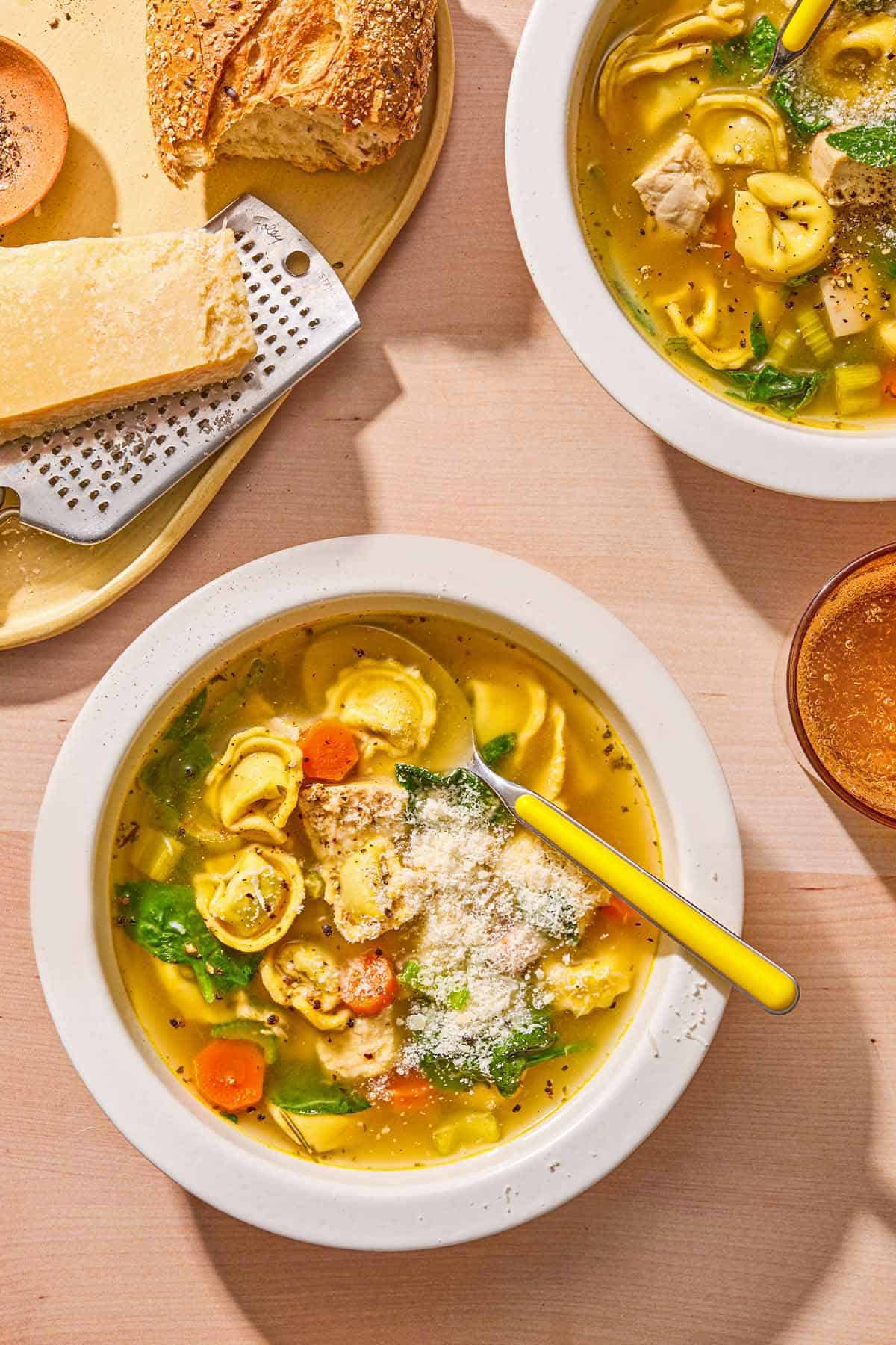 Chicken tortellini soup topped with grated Parmigiano Reggiano cheese in a bowl with a spoon. Next to this is another bowl of the soup with a spoon, a glass of sparkling water, and a plate with crusty bread, Parmigiano Reggiano cheese and a cheese grater.