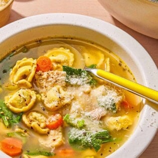 Chicken tortellini soup topped with grated Parmigiano Reggiano cheese in a bowl with a spoon. Next to this is a large pot with the rest of the soup and bowls of pepper and grated Parmigiano Reggiano cheese.