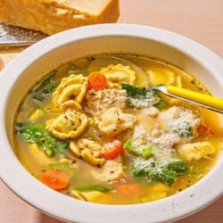 Chicken tortellini soup topped with grated Parmigiano Reggiano cheese in a bowl with a spoon. Next to this is a block of the Parmigiano Reggiano cheese, a cheese grater, and a small bowl of pepper.
