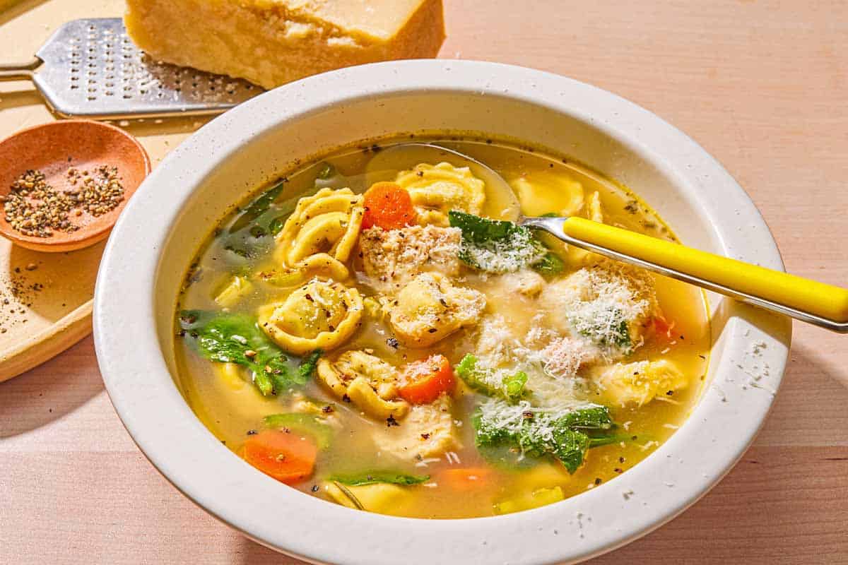 Chicken tortellini soup topped with grated Parmigiano Reggiano cheese in a bowl with a spoon. Next to this is a block of the Parmigiano Reggiano cheese, a cheese grater, and a small bowl of pepper.