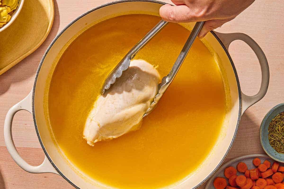 A chicken breast for the tortellini soup being added to the chicken broth with a pair of tongs. Next to this is a plate of sliced carrots and a bowl of Italian seasoning.