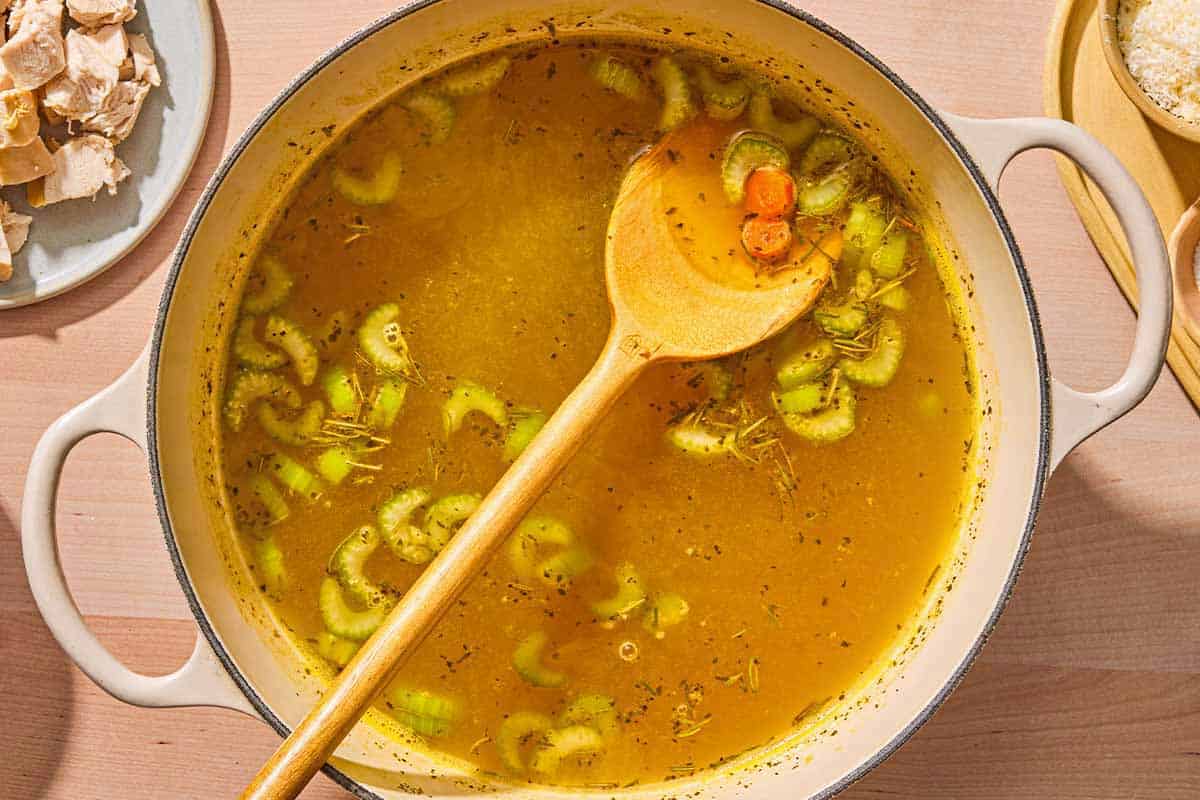 The vegetables for the tortellini soup combined with the broth in a large pot with a wooden spoon. Next to this is a plate of cooked chicken.