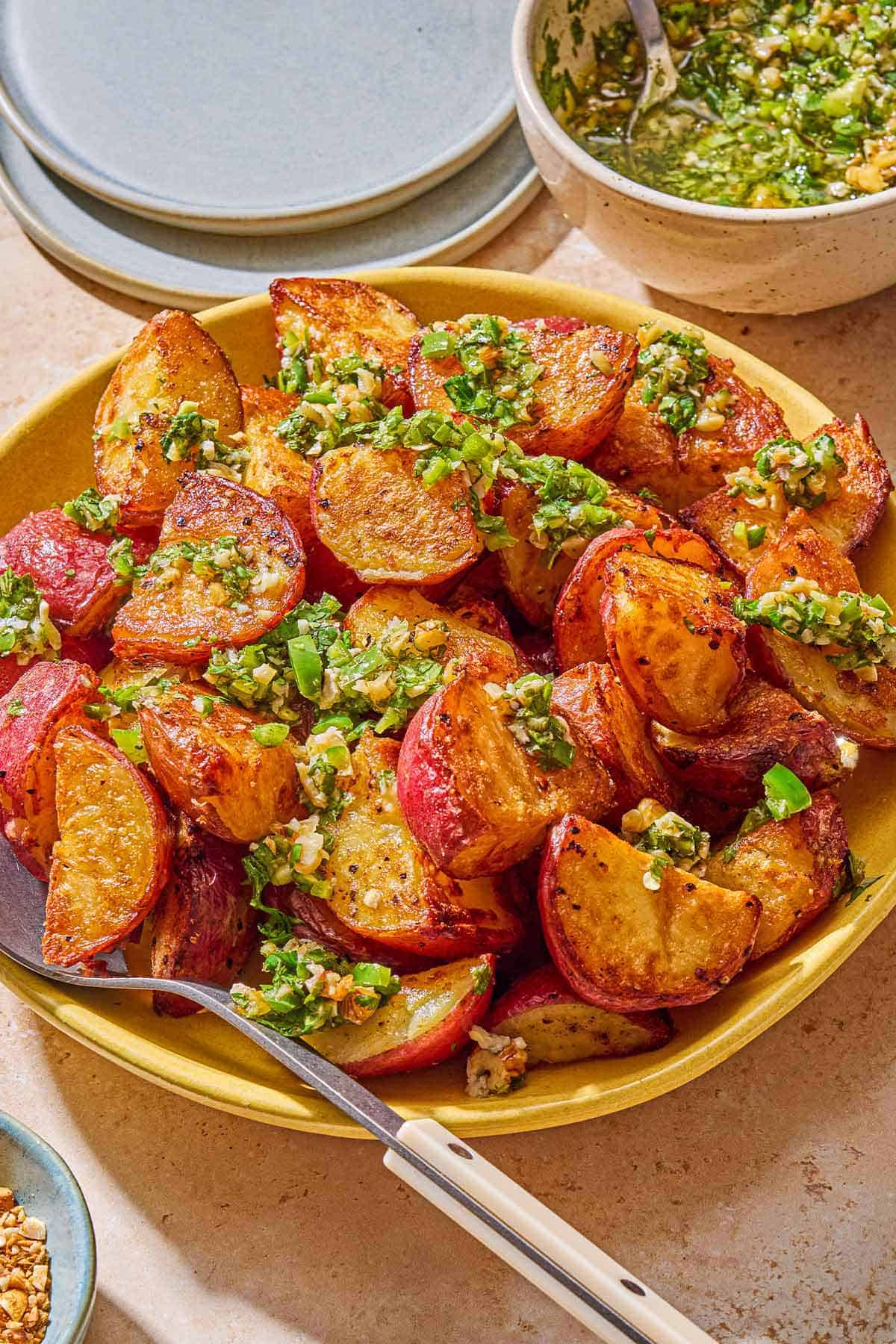 Crispy red skinned potatoes drizzled with walnut cilantro chili sauce in a bowl with a serving spoon. Next to this is a bowl of the sauce with a spoon and a stack of 2 plates.