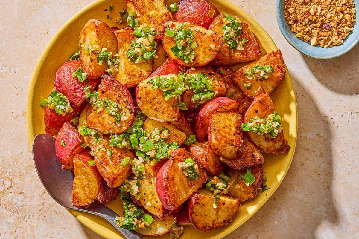 Crispy red skinned potatoes drizzled with walnut cilantro chili sauce in a bowl with a serving spoon. Next to this is a bowl of chopped walnuts.