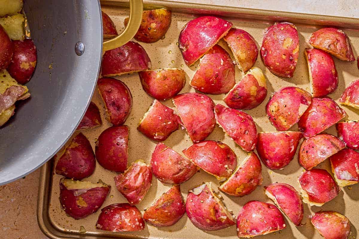 The seasoned, boiled red skinned potatoes spread evenly on a sheet pan. Next to this is a pot with more of the boiled potatoes.