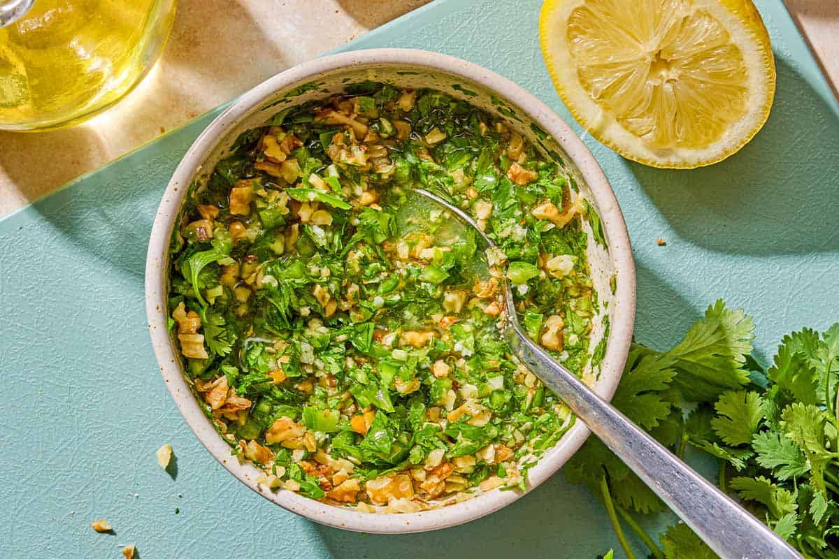 The walnut cilantro chili sauce in a bowl with a spoon next to a lemon half and cilantro on a cutting board.