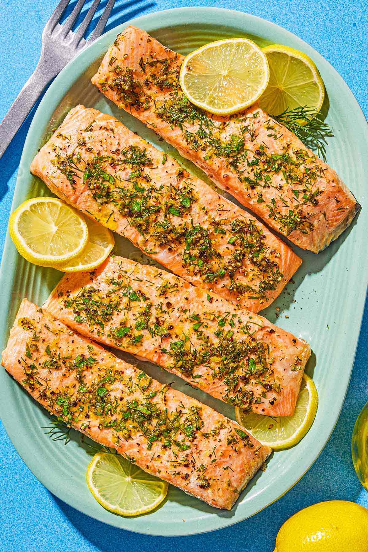 Four cooked lemon pepper salmon fillets on a platter with lemon wheels. Next to this is a lemon and a serving fork.