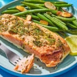 A lemon pepper salmon fillet on a plate with roasted green beans, a lemon wheel, and a fork containing a bit of the salmon.
