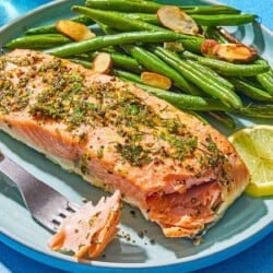A lemon pepper salmon fillet on a plate with roasted green beans, a lemon wheel, and a fork containing a bit of the salmon.