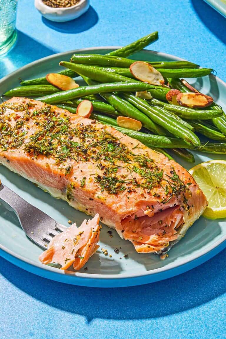 A lemon pepper salmon fillet on a plate with roasted green beans, a lemon wheel, and a fork containing a bit of the salmon.