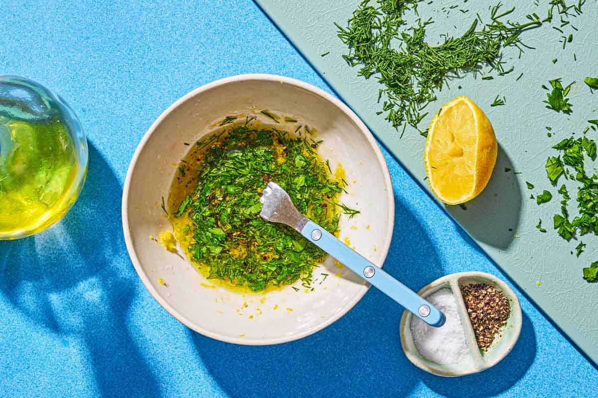 The lemon pepper sauce in a bowl with a fork. Next to this is a bottle of olive oil, a bowl of salt and pepper, and a juiced lemon, some fresh dill and some fresh parsley on a cutting board.