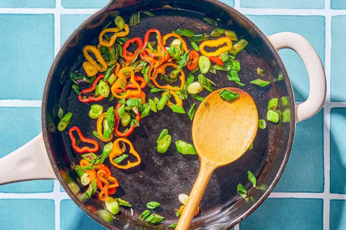 Baby bell pepper rings and chopped green onions being sautéed in a skillet with a wooden spoon.