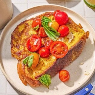 A slice of the savory french toast with the veggie topping on a plate. Next to this is a knife and fork, a small bowl Parmigiano Reggiano cheese, and a cup of orange juice.