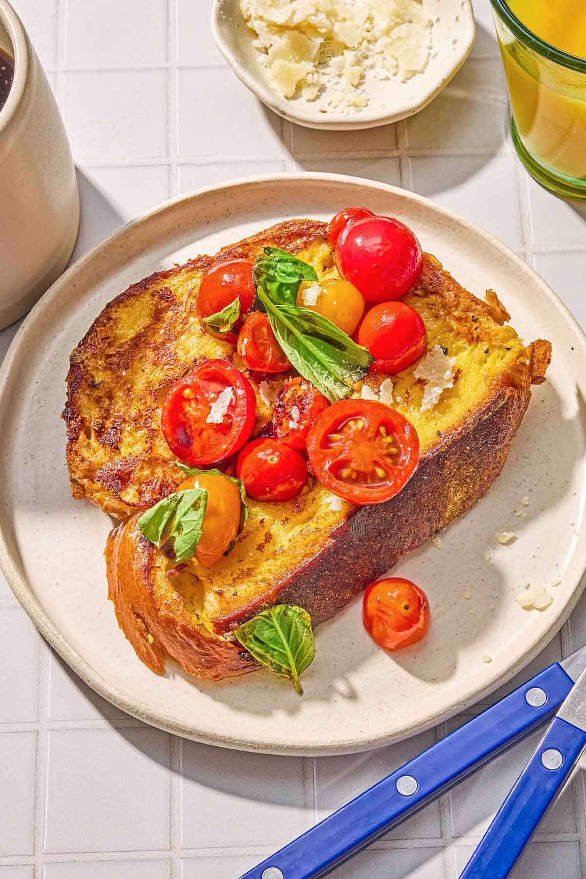 A slice of the savory french toast with the veggie topping on a plate. Next to this is a knife and fork, a small bowl Parmigiano Reggiano cheese, and a cup of orange juice.