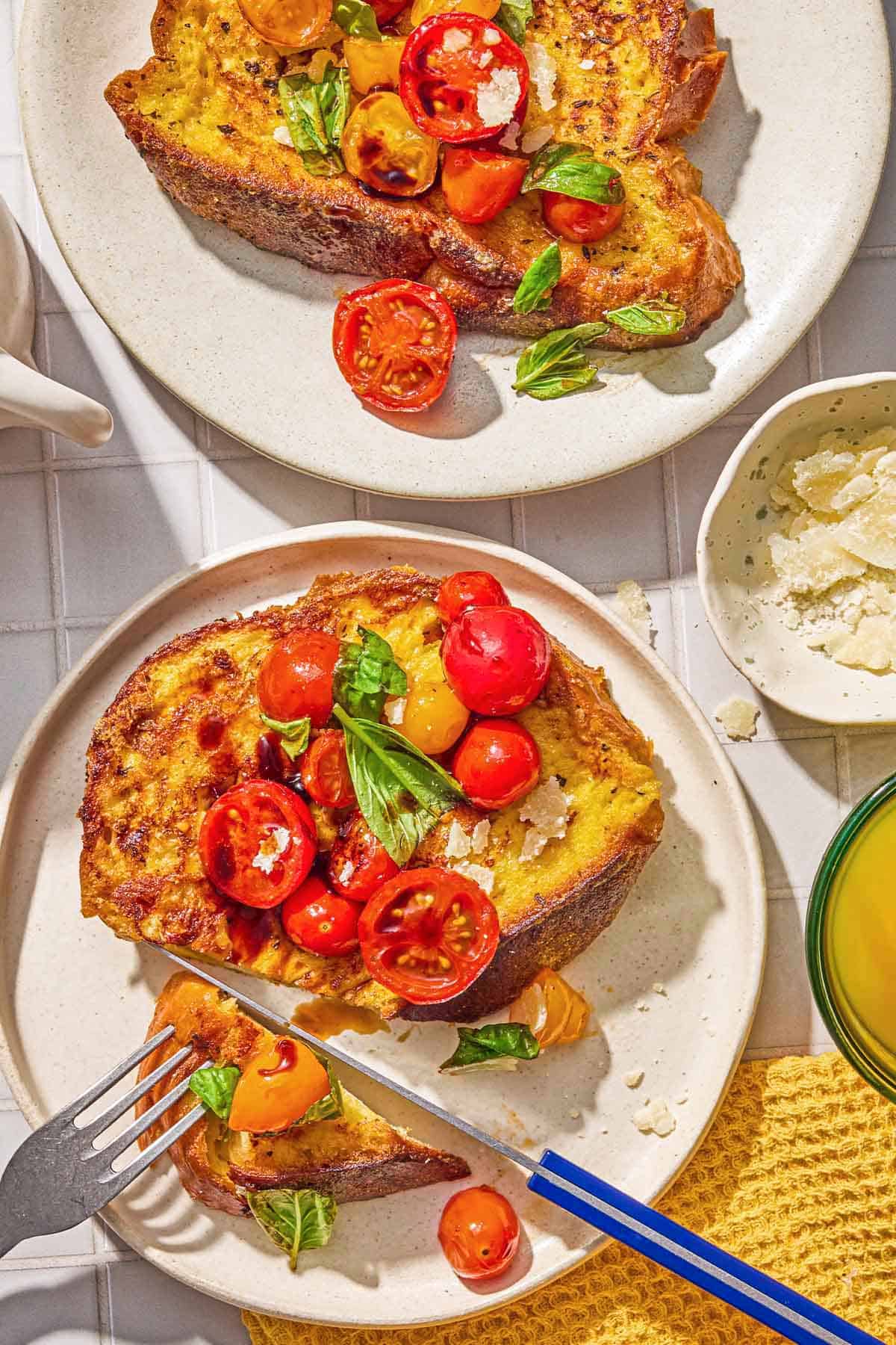 A slice of the savory french toast on a plate being sliced with a knife and fork. Next to this is another plate with piece of the french toast and a small bowl Parmigiano Reggiano cheese.