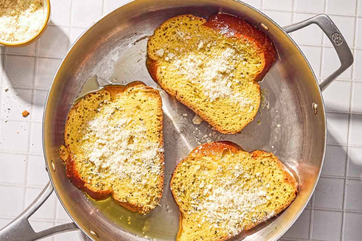 Three slices of the savory french toast topped with finely grated Parmigiano Reggiano cheese cooking in a skillet. Next to this is a bowl of the grated cheese.
