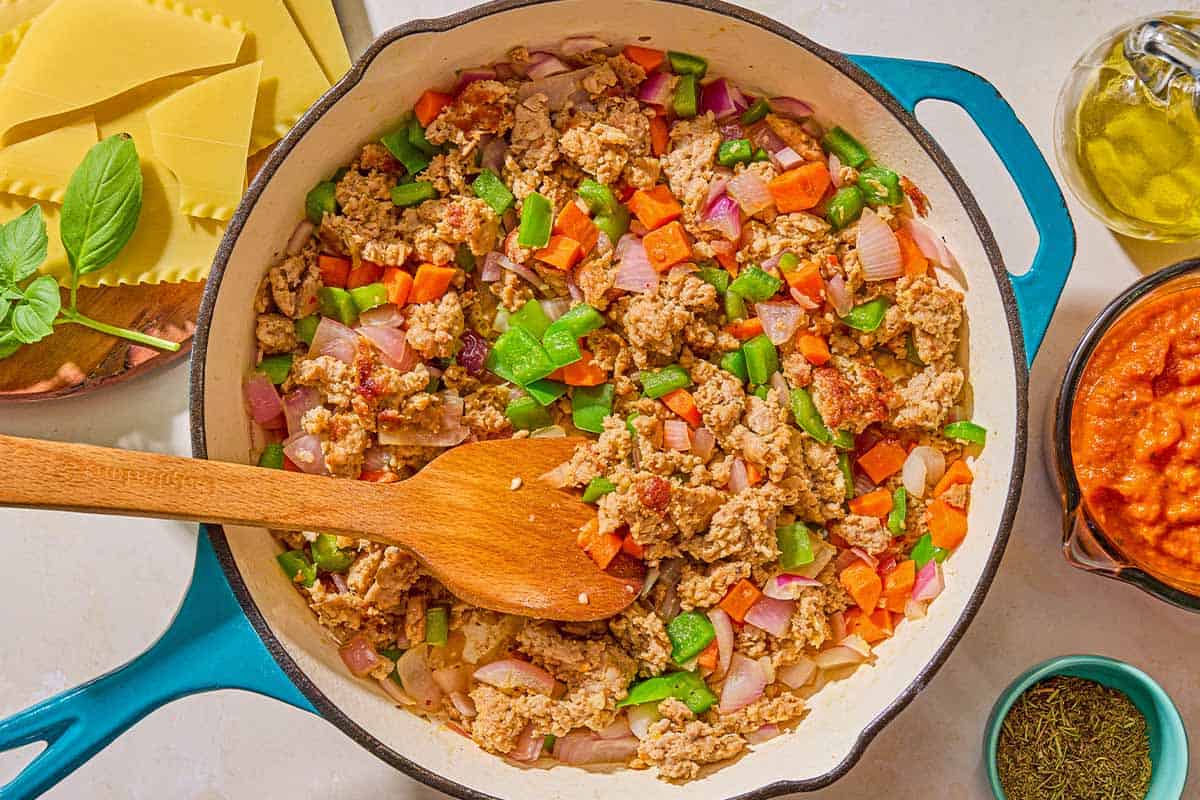 The sausage and vegetables for the skillet lasagna being sautéed in a skillet with a wooden spoon. Next to this is a bowl of Italian seasoning, a measuring cup with marinara, a bottle of olive oil, the uncooked lasagna noodles, and a sprig of fresh basil.