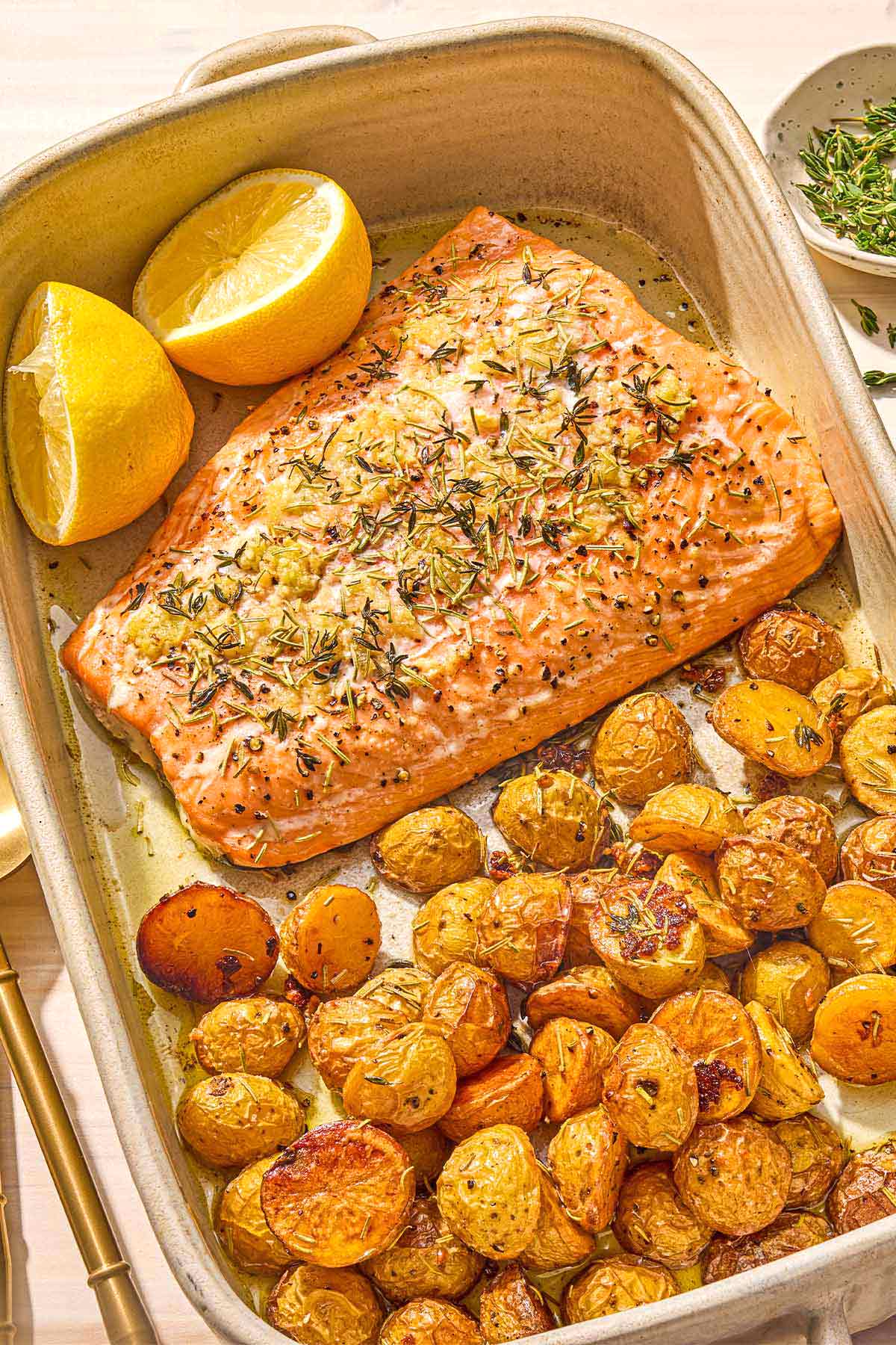 The slow roasted salmon and potatoes in a baking dish with 2 lemon halves. Next to this is a bowl small bowl of chopped thyme.