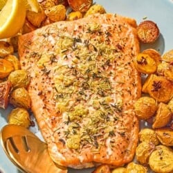 The slow roasted salmon and potatoes on a platter with lemon wedges and serving utensils. Next to this is a bowl small bowl of chopped thyme.